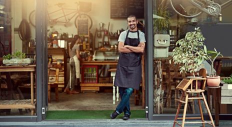 Bilde av en mann med skjegg som står med armene i kors i døren til en kaffebar med forkle på seg