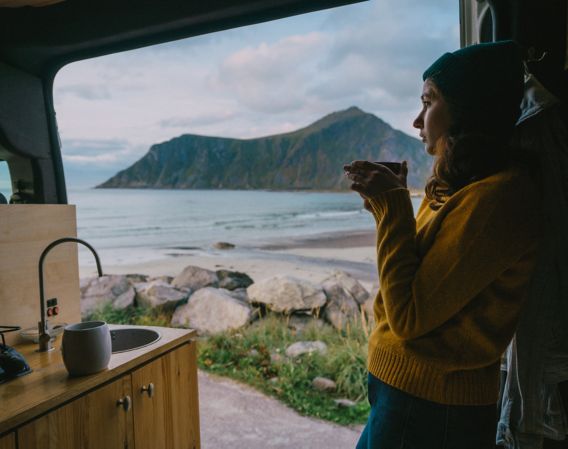 Bilde av kvinne som står inni en bobil med kaffekopp i hånden og fin utsikt i bakgrunnen