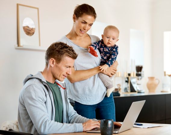 Man sitting at a table and looking at a computer, woman standing by the side of the table and holding a child while looking at a computer
