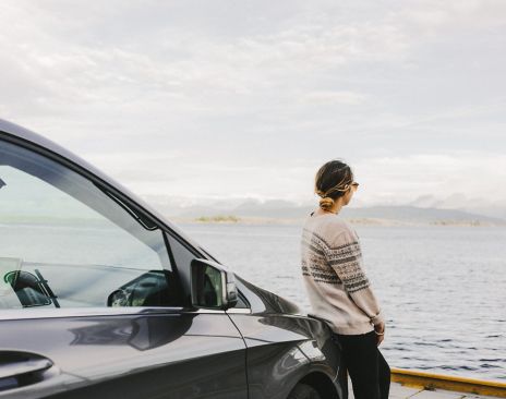 Ung dame ser utover fjorden mens hun lener seg på bilen sin.
